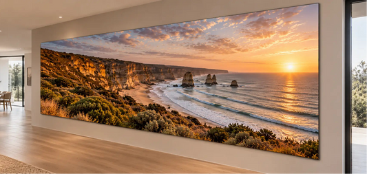 A large panoramic wall art print showing a coastal cliff and sea stacks at sunset, displayed in a modern interior hallway with wood flooring and glass doors.
